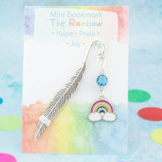 A rainbow charm bookmark. The metal bookmark is in the shape of a feather and it has a crystal connector between it and the charm. The bookmark is backed onto a card with the symbolic meanings of the rainbow - hope, pride and joy.