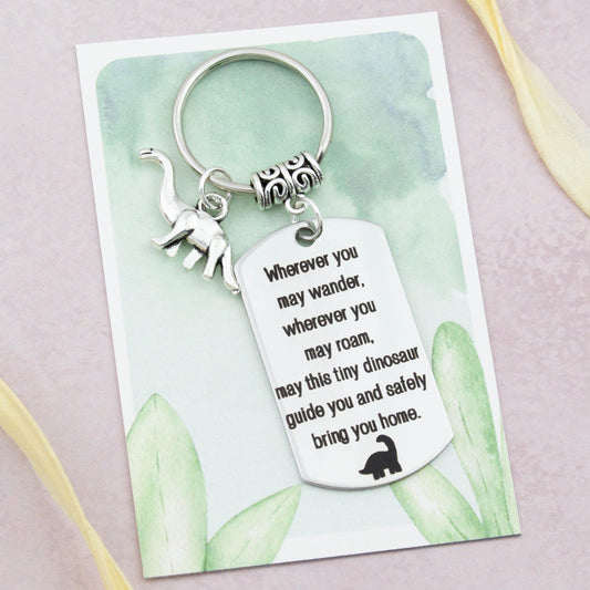 A keyring with two charms on a matching backing card. The charms are a silver Dinosaur and a quote charm that reads “Wherever you may wander, wherever you may roam, may this tiny Dinosaur guide you and bring you safely home”