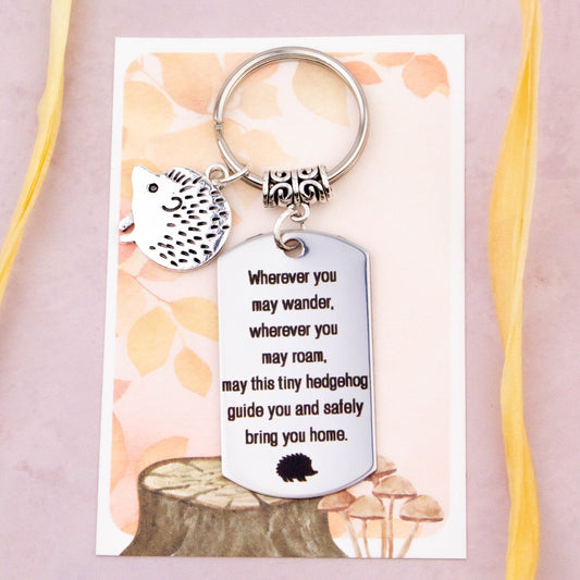 A keyring with two charms on a matching backing card. The charms are a silver hedgehog and a quote charm that reads “Wherever you may wander, wherever you may roam, may this tiny hedgehog guide you and bring you safely home”