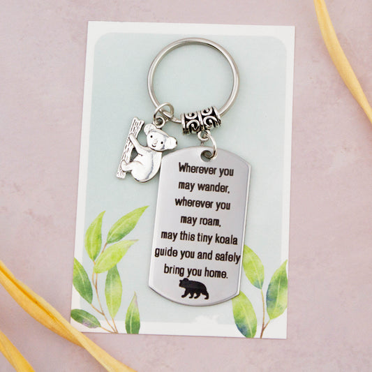 A keyring with two charms on a matching backing card. The charms are a silver koala and a quote charm that reads “Wherever you may wander, wherever you may roam, may this tiny koala guide you and bring you safely home”