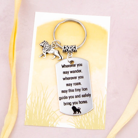 A keyring with two charms on a matching backing card. The charms are a silver lion and a quote charm that reads “Wherever you may wander, wherever you may roam, may this tiny lion guide you and bring you safely home”