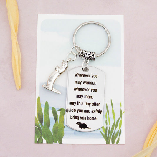 A keyring with two charms on a matching backing card. The charms are a silver otter and a quote charm that reads “Wherever you may wander, wherever you may roam, may this tiny otter guide you and bring you safely home”