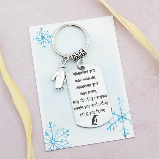 A keyring with two charms on a matching backing card. The charms are a silver Penguin and a quote charm that reads “Wherever you may wander, wherever you may roam, may this tiny Penguin guide you and bring you safely home”
