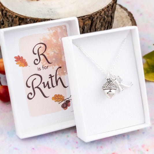 A silver chain necklace with an acorn pendant and initial charm. The gift box is personalised with the name of the recipient.