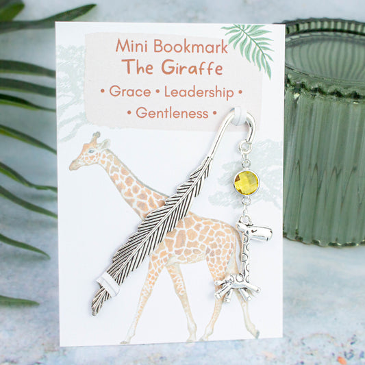 A giraffe charm hanging from a metal bookmark. It has a yellow crystal. The bookmark is backed onto a card with what the charm means symbolically.