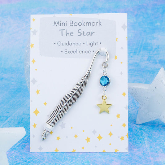 A gold star charm hanging from a metal bookmark. It has a blue crystal. The bookmark is backed onto a card with what the charm means symbolically.