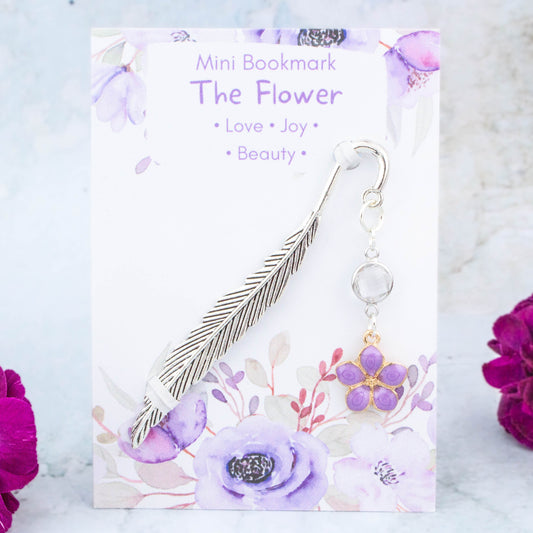 A purple flower charm bookmark. The metal bookmark is in the shape of a feather and it has a crystal connector between it and the charm. The bookmark is backed onto a card with the symbolic meanings of the flower. Love, joy and beauty.