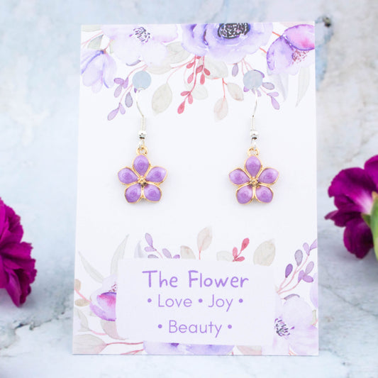 A pair of purple flower charm earrings backed onto a card with the symbolic meanings of a flower. "Love, Joy and Beauty"