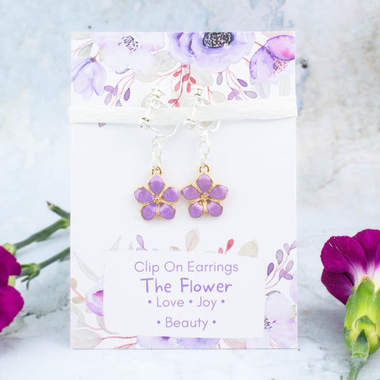 A pair of purple flower clip on earrings. It's backed onto a card that says the symbolic meanings of the flower - love, joy and beauty.