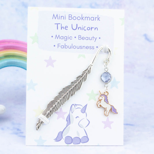 A purple unicorn charm hanging from a metal bookmark. It has a purplecrystal. The bookmark is backed onto a card with what the charm means symbolically.