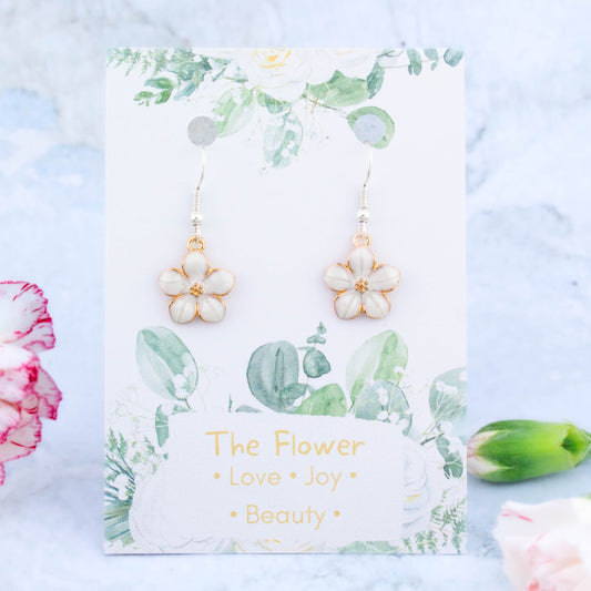 A pair of white flower charm earrings backed onto a card with the symbolic meanings of a flower. "Love, Joy and Beauty"