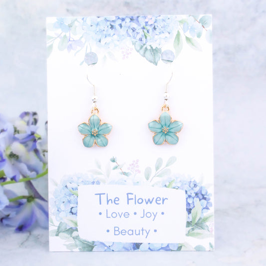 A pair of blue flower charm earrings backed onto a card with the symbolic meanings of a flower. "Love, Joy and Beauty"