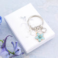 A blue flower charm keychain personalised with an initial and birthstone sitting on a white gift box silver foiled with the logo.