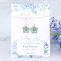 A pair of blue flower clip on earrings. It's backed onto a card that says the symbolic meanings of the flower - love, joy and beauty.