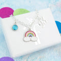 A silver enamel rainbow charm bracelet with an initial and birthstone charm. It sits on a white gift box with a silver foil logo on the lid.