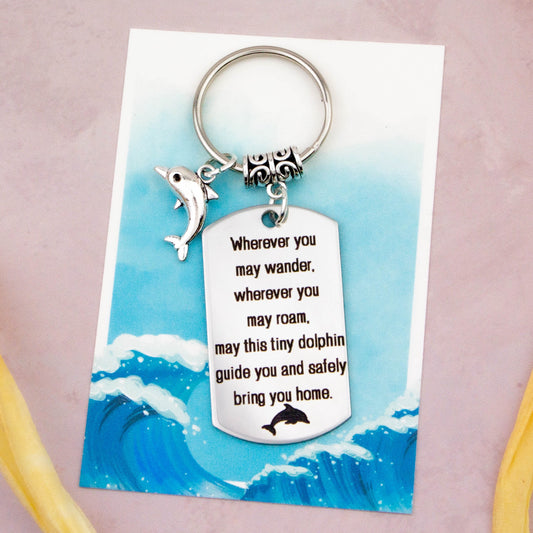 A keyring with two charms on a matching backing card. The charms are a silver dolphin and a quote charm that reads “Wherever you may wander, wherever you may roam, may this tiny dolphin guide you and bring you safely home” 