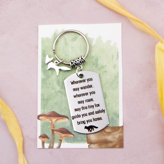 A keyring with two charms on a matching backing card. The charms are a silver fox and a quote charm that reads “Wherever you may wander, wherever you may roam, may this tiny fox guide you and bring you safely home” 