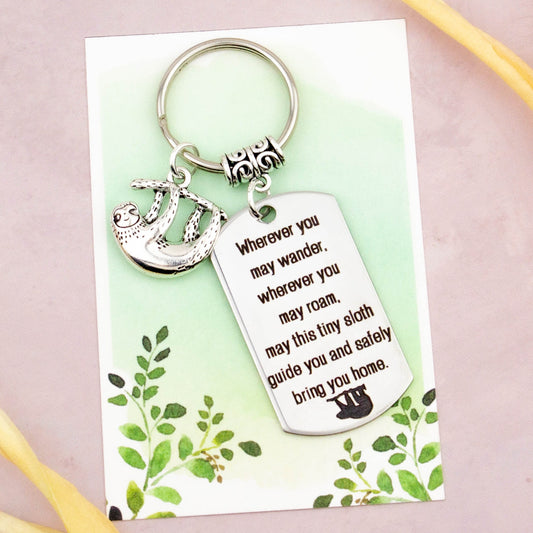 A keyring with two charms on a matching backing card. The charms are a silver sloth and a quote charm that reads “Wherever you may wander, wherever you may roam, may this tiny sloth guide you and bring you safely home” 