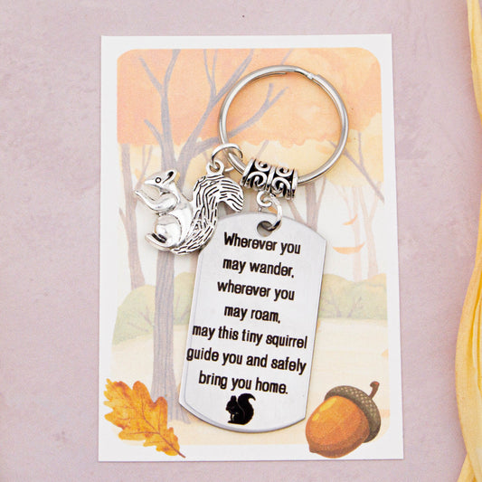 A keyring with two charms on a matching backing card. The charms are a silver squirrel and a quote charm that reads “Wherever you may wander, wherever you may roam, may this tiny squirrel guide you and bring you safely home” 