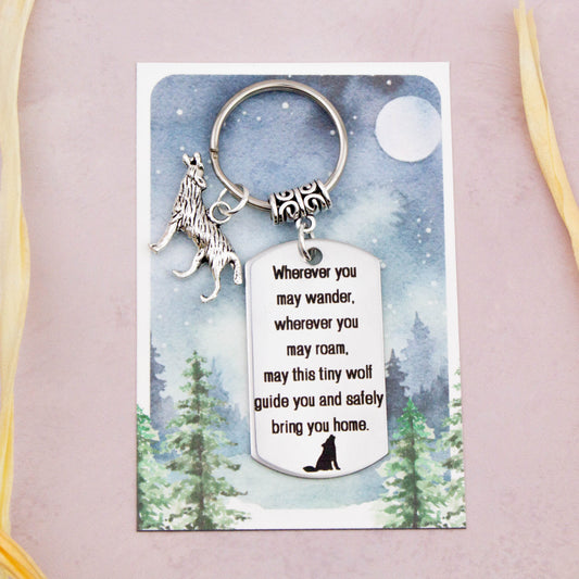 A keyring with two charms on a matching backing card. The charms are a silver wolf and a quote charm that reads “Wherever you may wander, wherever you may roam, may this tiny wolf guide you and bring you safely home” 
