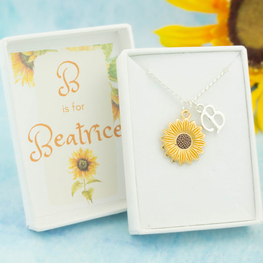 A silver chain necklace with a yellow enamel sunflower pendant and initial charm. The gift box is personalised with the name of the recipient. 