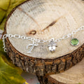 A silver 3D acorn charm bracelet with an initial and birthstone charm. It sits on a white gift box with a silver foil logo on the lid.