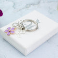 A purple flower charm keychain personalised with an initial and birthstone sitting on a white gift box silver foiled with the logo.