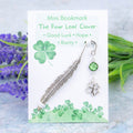 A four leaf clover charm hanging from a metal bookmark. It has a green crystal. The bookmark is backed onto a card with what the charm means symbolically.  