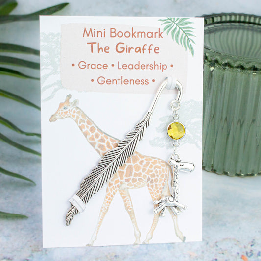 A giraffe charm hanging from a metal bookmark. It has a yellow crystal. The bookmark is backed onto a card with what the charm means symbolically.  