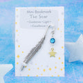 A gold star charm hanging from a metal bookmark. It has a blue crystal. The bookmark is backed onto a card with what the charm means symbolically.  