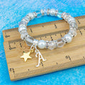 A silver and white gold star and silver initial charm bracelet against a ruler. 