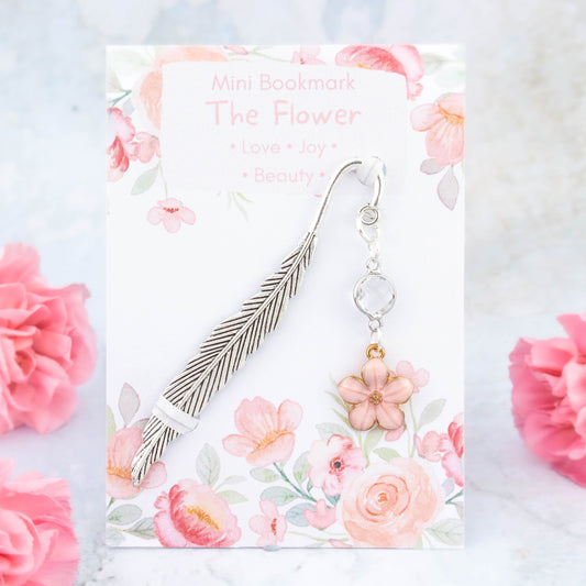 A pink flower charm bookmark. The metal bookmark is in the shape of a feather and it has a crystal connector between it and the charm. The bookmark is backed onto a card with the symbolic meanings of the flower. Love, joy and beauty.