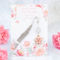 A pink flower charm bookmark. The metal bookmark is in the shape of a feather and it has a crystal connector between it and the charm. The bookmark is backed onto a card with the symbolic meanings of the flower. Love, joy and beauty.