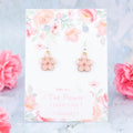 A pair of pink flower charm earrings backed onto a card with the symbolic meanings of a flower. 