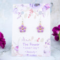 A pair of purple flower charm earrings backed onto a card with the symbolic meanings of a flower. 