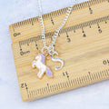 A purple and white enamel unicorn and initial charm necklace against a ruler. 