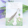 A white rabbit charm hanging from a metal bookmark. It has a green crystal. The bookmark is backed onto a card with what the charm means symbolically.  