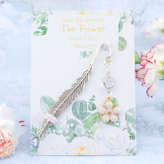 A white flower charm bookmark. The metal bookmark is in the shape of a feather and it has a crystal connector between it and the charm. The bookmark is backed onto a card with the symbolic meanings of the flower. Love, joy and beauty.