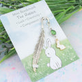 A white rabbit charm on a metal bookmark in the shape of a feather. It is backed onto a card with the symbolic meanings of the charm. 